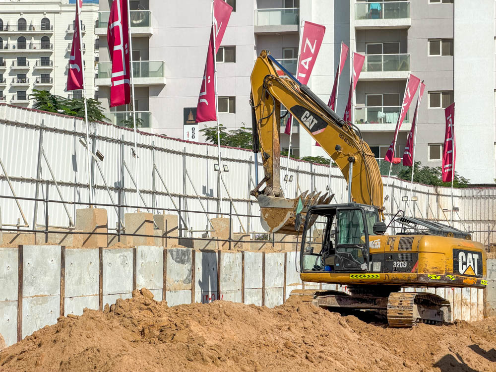 Cove Grand By Imtiaz A yellow excavator is parked on a construction site with sand, walls, and colorful flags in the background.